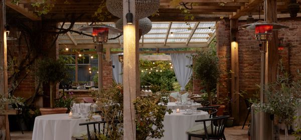 Restaurant tables in a courtyard setting under a wooden pergola with climbing plants.